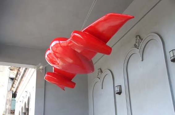 Sculpture of a glossy red bat-winged form suspended from a ceiling against an arched stone wall.