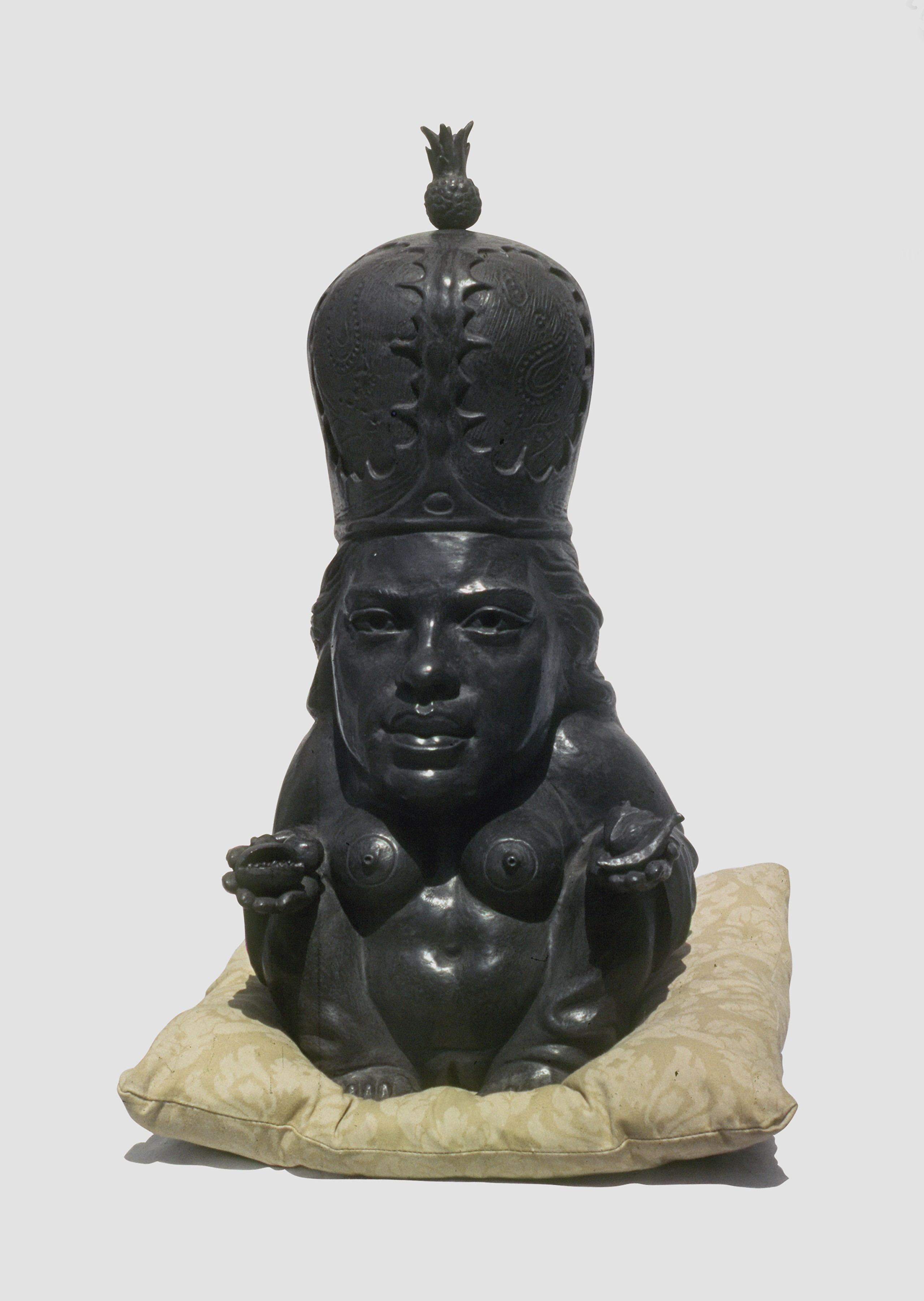 Dark bronze sculpture of a bare-chested female figure wearing a tall crown-like headdress topped with a pineapple finial, kneeling on a cushion with hands cupped outward.