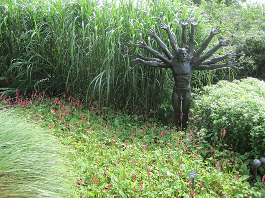 The full bronze many-armed figure sculpture standing amid tall grasses and pink flowers in a garden.