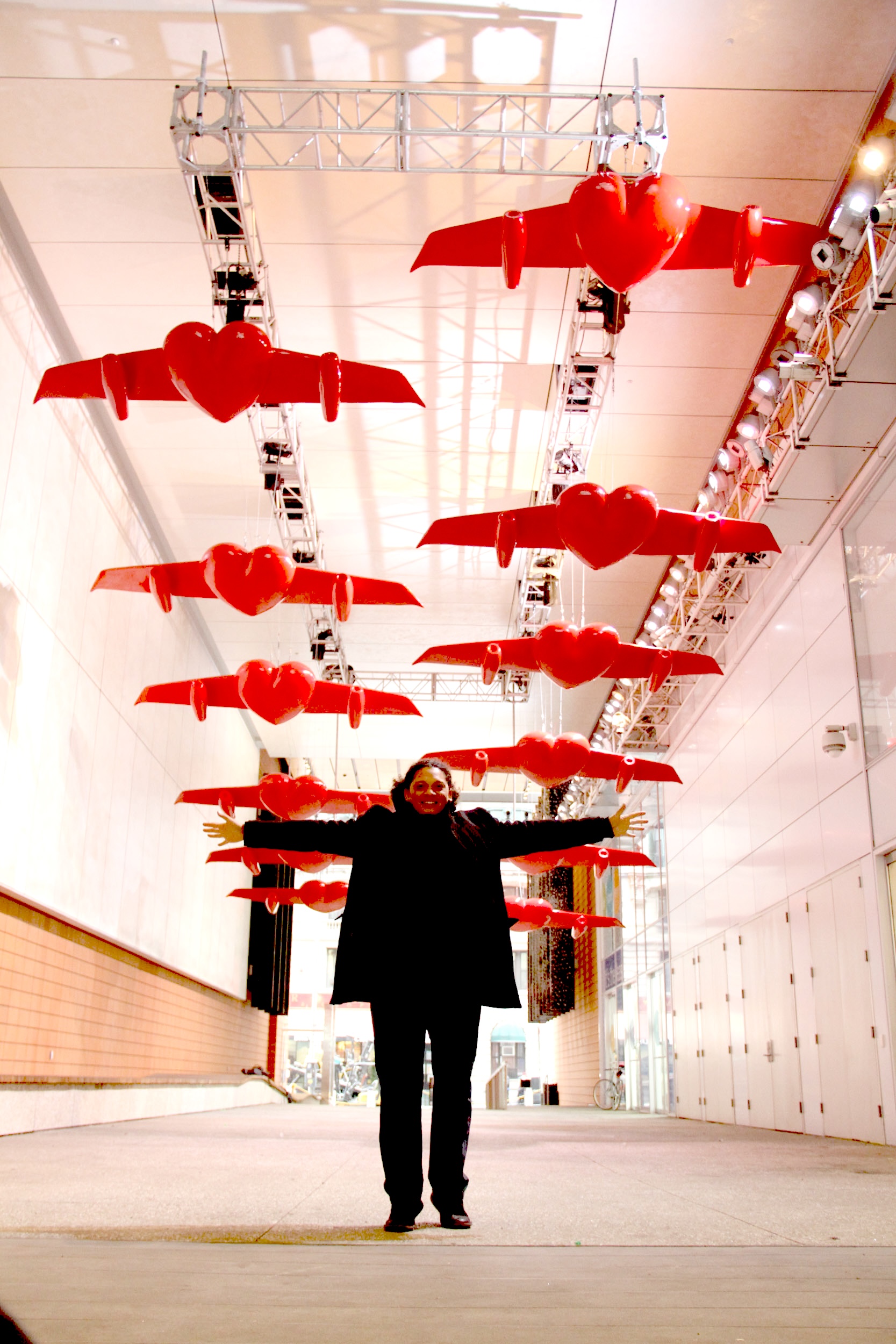 Photograph of a woman standing with arms outstretched beneath rows of glossy red heart-shaped sculptures fitted with wings, suspended from ceiling trusses in an indoor walkway.