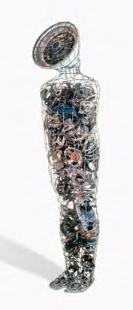 A standing wire-mesh human silhouette with a round gauge-like head, its body densely packed with tangled gears, dials, and salvaged mechanical parts visible through the mesh.