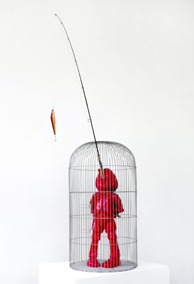 A red Pinocchio-like figure enclosed in a birdcage, its long nose reaching out through the top holding a fishing line.