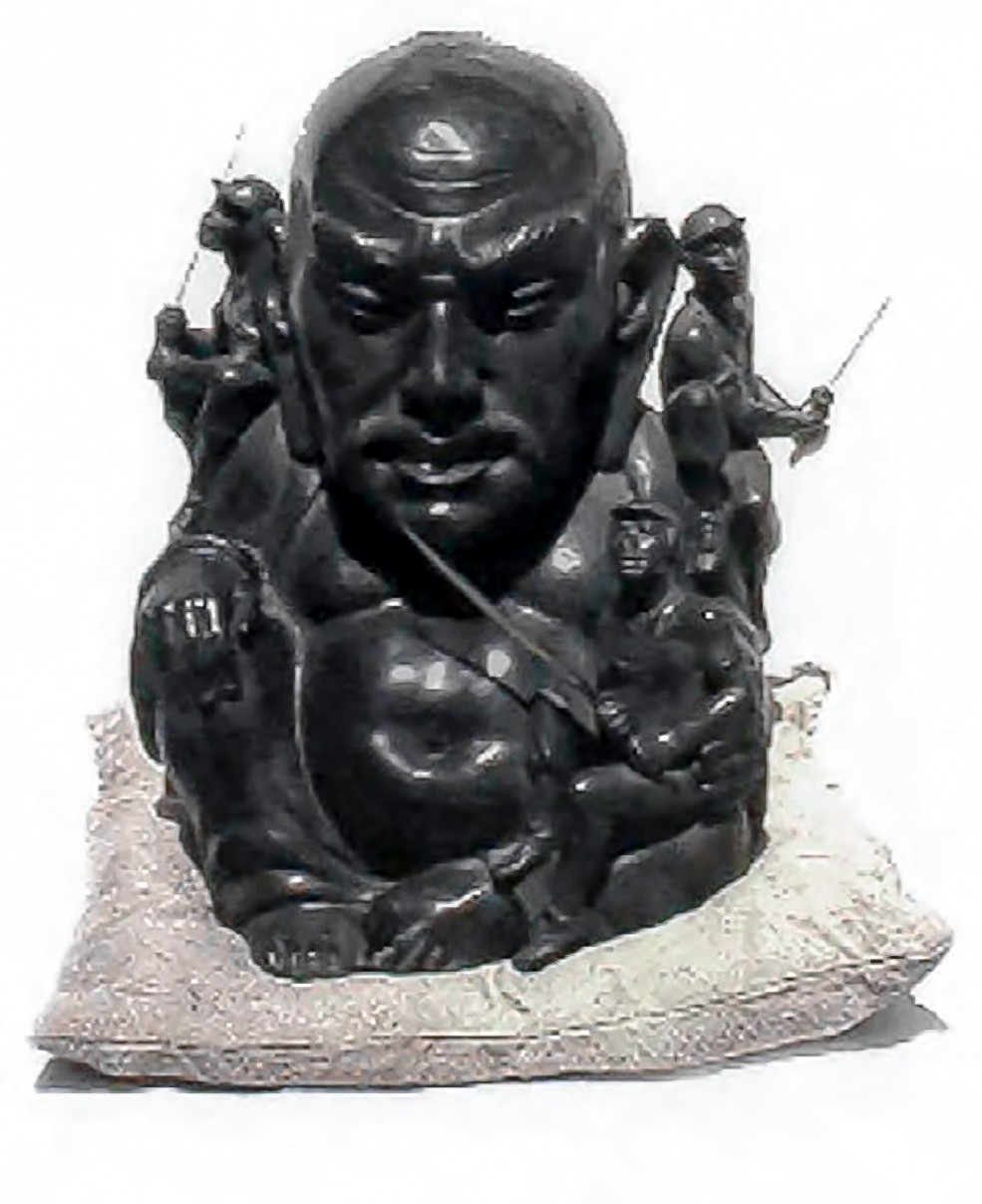 Bronze sculpture of a bald head with an eye carved into the forehead, seated on a cushion, surrounded by small figures climbing and swinging around its shoulders and arms.