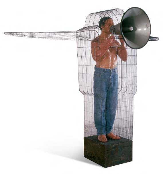 Sculpture of a man's torso and legs encased in a wire-mesh cage, holding a large megaphone up to his mouth, standing on a dark wooden pedestal.
