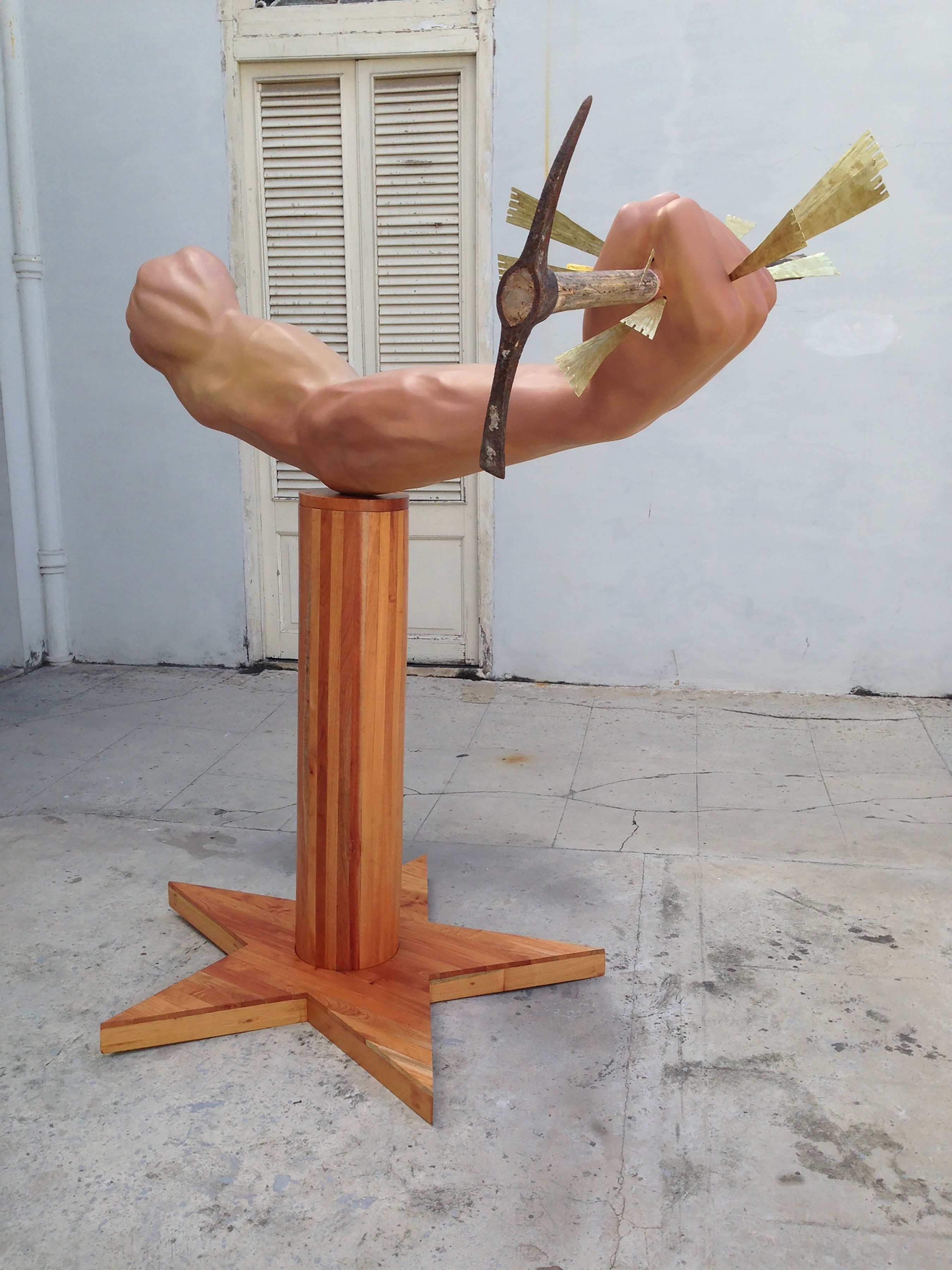 Sculpture of a muscular forearm and clenched fist mounted on a star-shaped wooden pedestal, gripping a rusted anchor-like tool pierced with golden arrow-shaped blades.