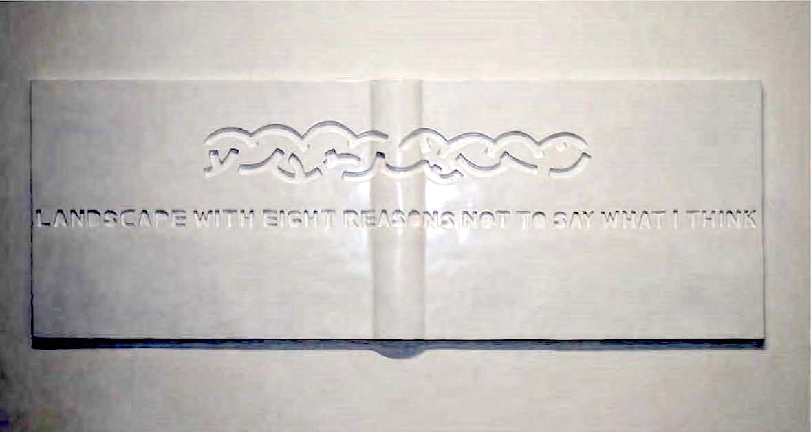 White relief sculpture shaped like an open book with a carved braided vine border across the top and the English phrase 'LANDSCAPE WITH EIGHT REASONS NOT TO SAY WHAT I THINK' cut into the surface.