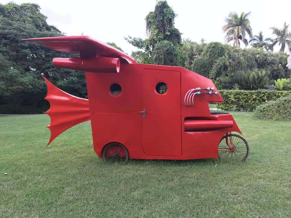 Outdoor sculpture of a red camper-van-like vehicle body fitted with a swept-back wing, a bat-wing tail fin, and bicycle wheels, parked on a lawn.