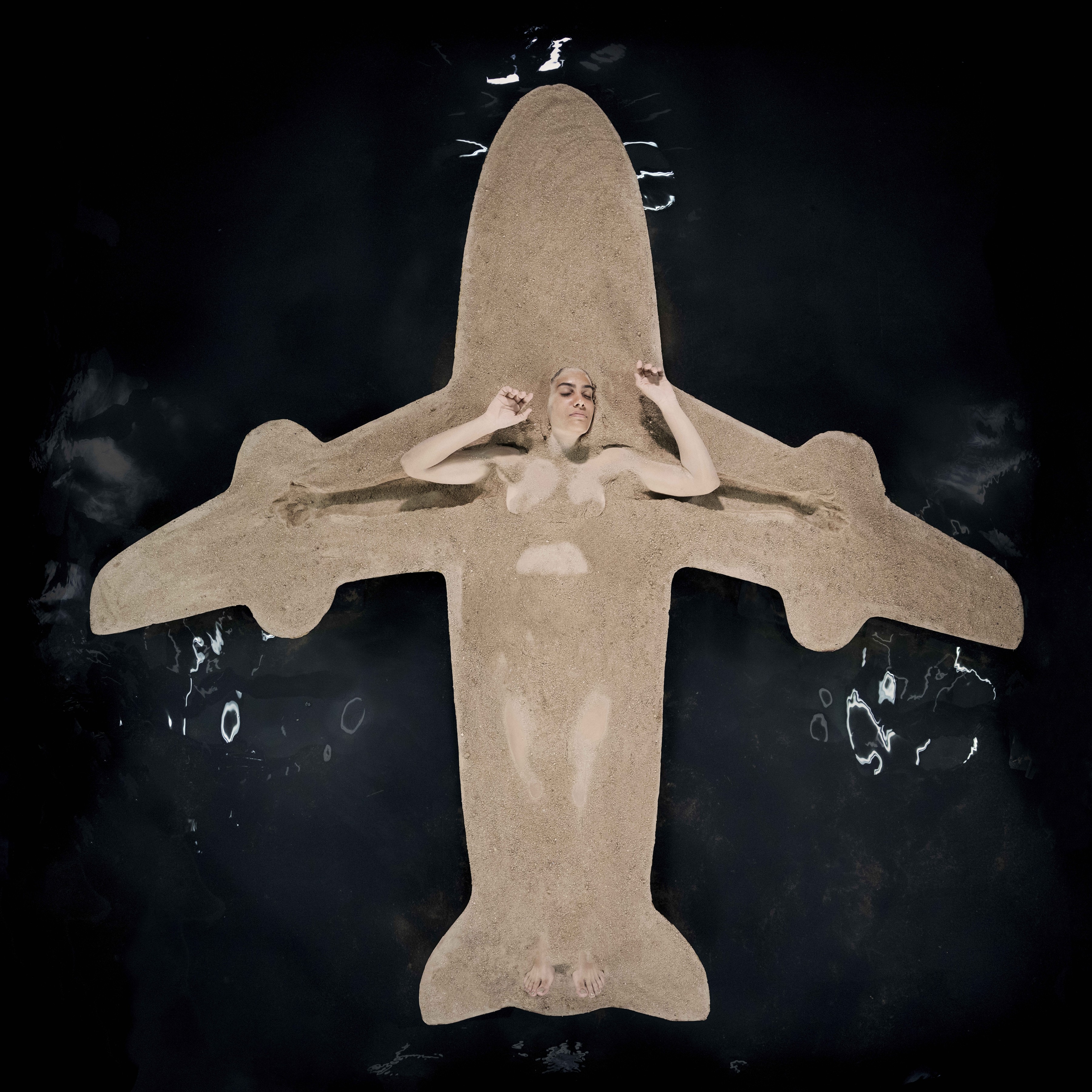 Overhead photograph of a nude woman lying in wet sand shaped into the silhouette of a jetliner, her body forming the fuselage and her raised arms tucked into the nose.