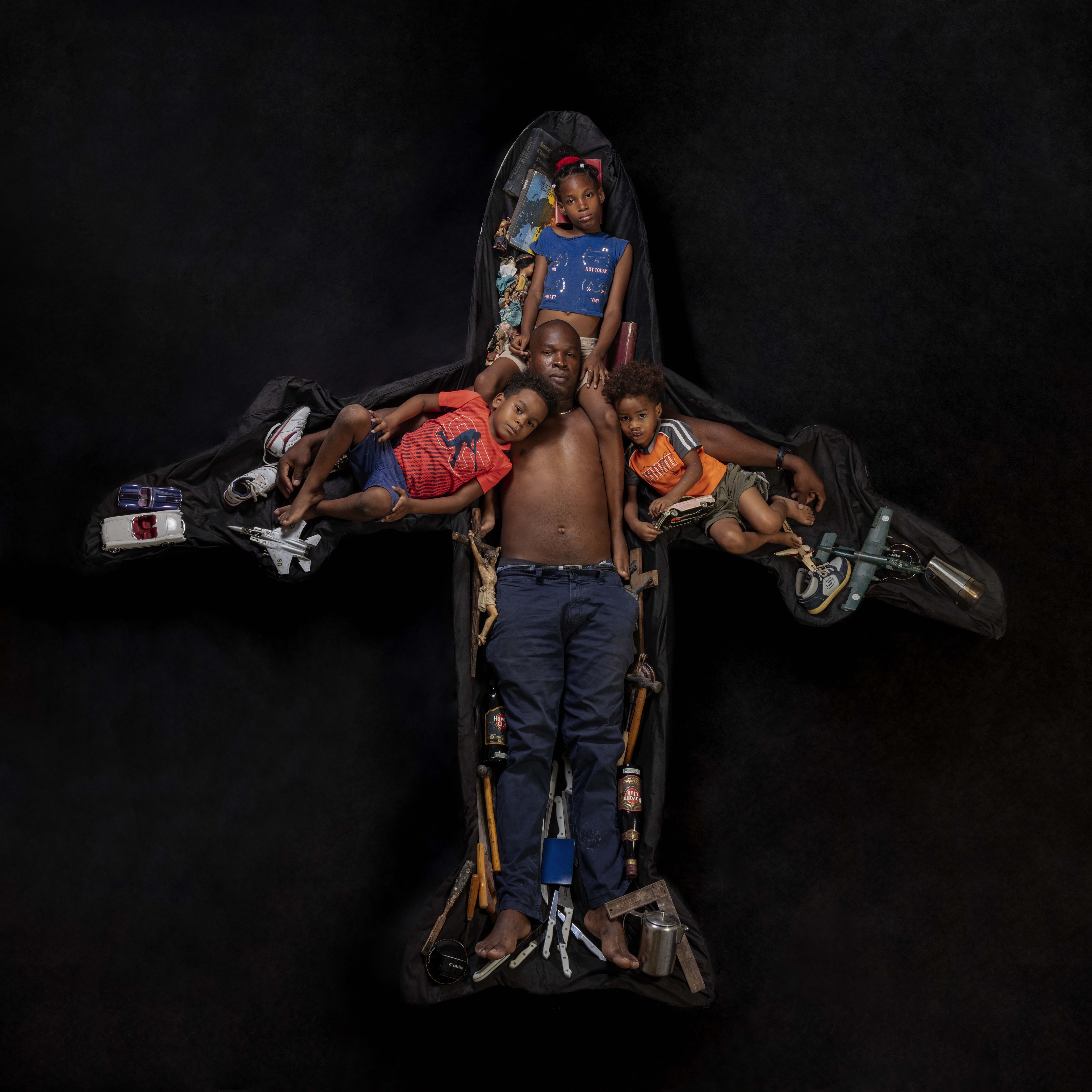Overhead photograph of a shirtless man and three children arranged inside a black fabric jetliner silhouette, surrounded by toy cars, a crucifix, tools, and a bottle of rum tucked along the wings and fuselage.