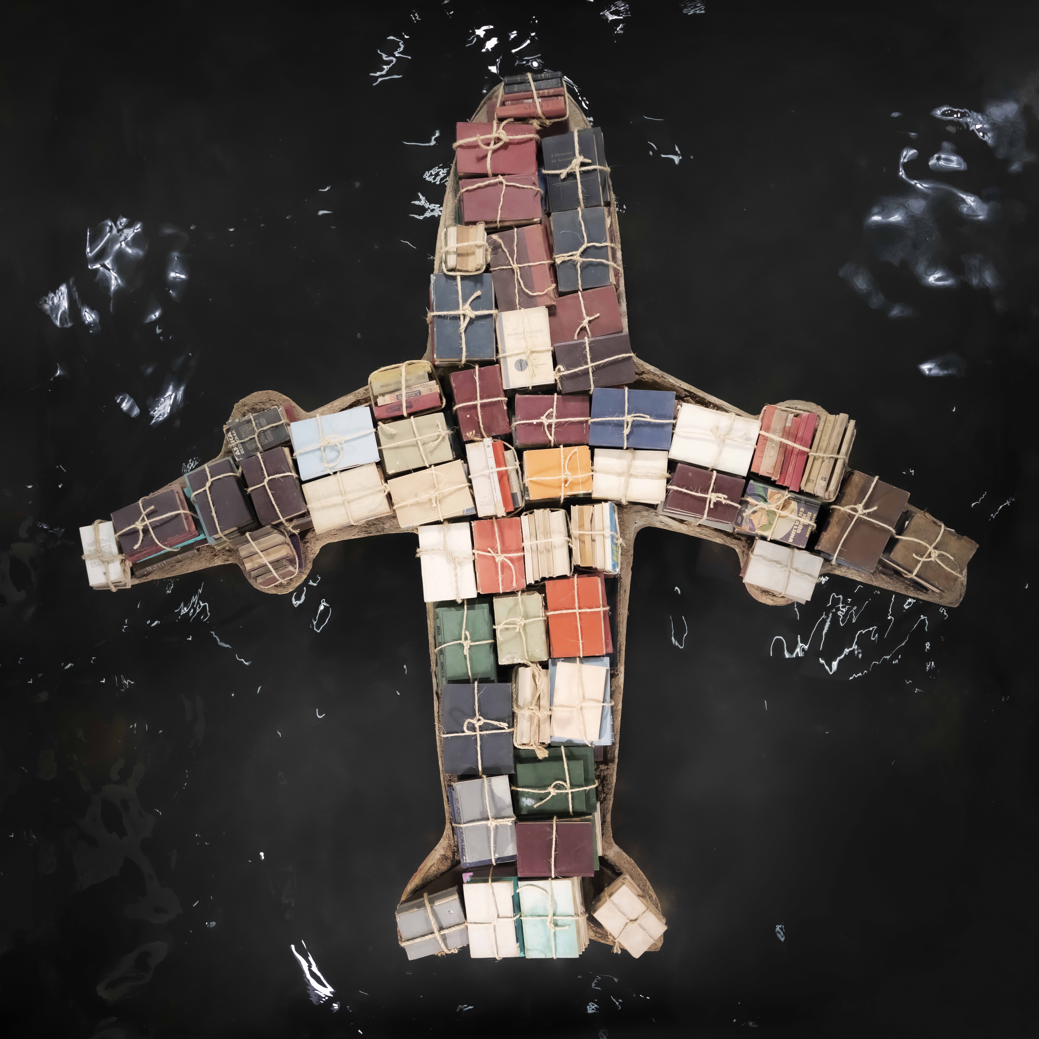 Overhead photograph of a jetliner silhouette formed from dozens of old books bundled with twine, their colored covers filling the fuselage and wings.
