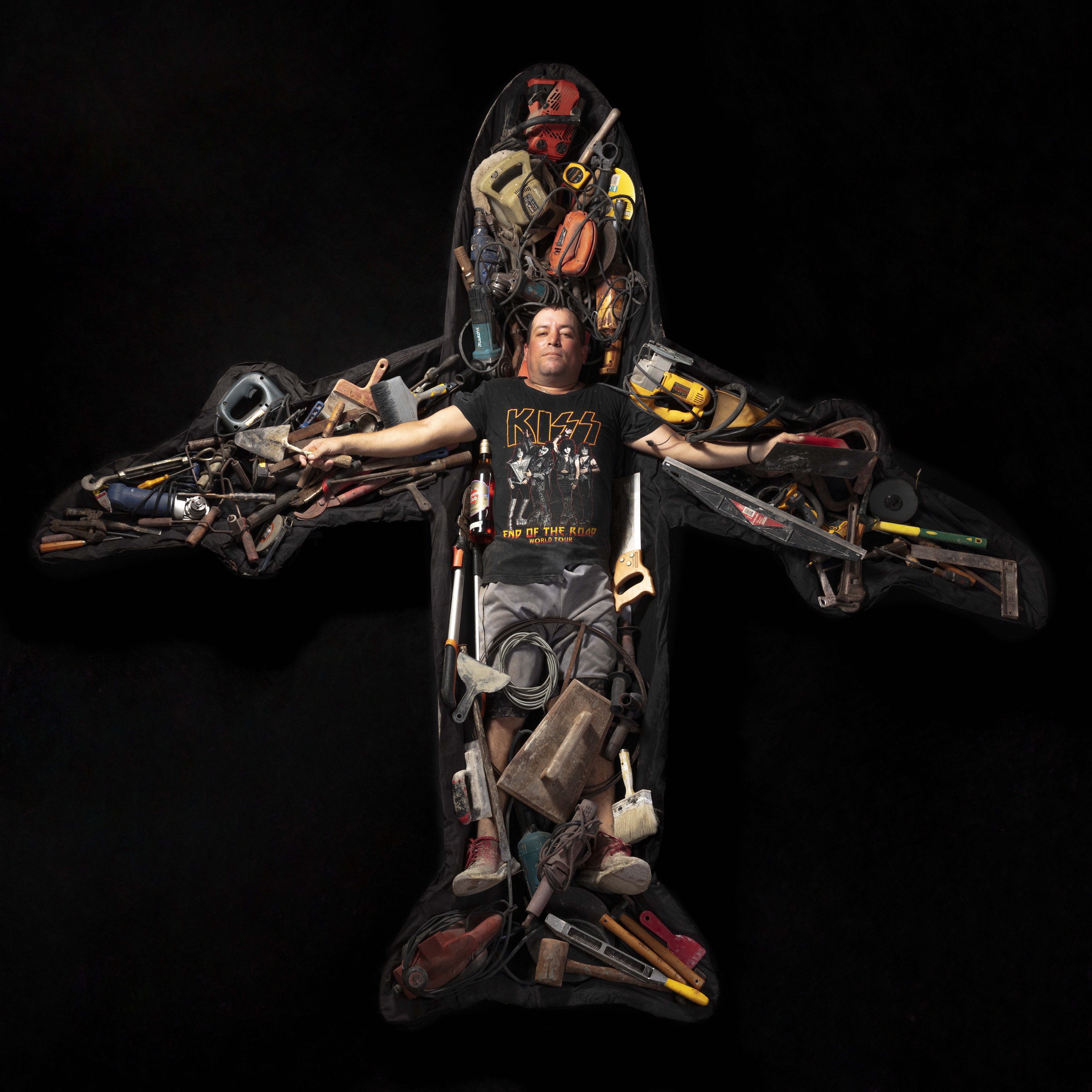 Overhead photograph of a man in a black KISS band T-shirt lying within a jetliner silhouette built entirely from hand tools and power tools, arms outstretched along the wings.
